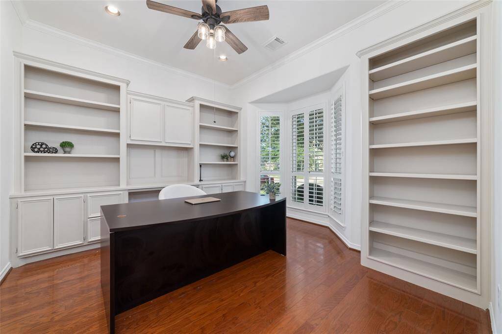 A bright home office with built-in bookshelves, a large desk, and a bay window with plantation shutters. The room features hardwood floors and a ceiling fan, offering a spacious and functional workspace.
