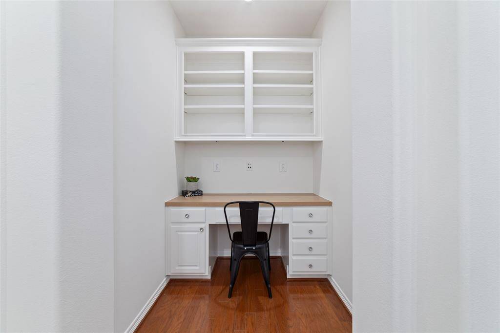 This nook with a built-in desk, white cabinetry, and open shelving is a perfect work space for getting school work done or a second office. The space is bright with a minimalist design, hardwood flooring, and a single chair, perfect for focused work or study.  It is located just off the 2 secondary bedrooms.