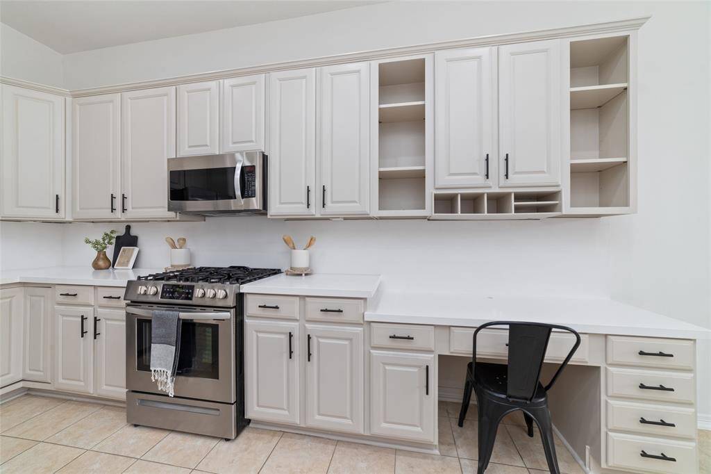 This kitchen features sleek, light-colored cabinetry with ample storage, a modern stainless steel stove and microwave, and a built-in desk area. The clean design is complemented by tiled flooring and neutral countertops, making it both functional and stylish.