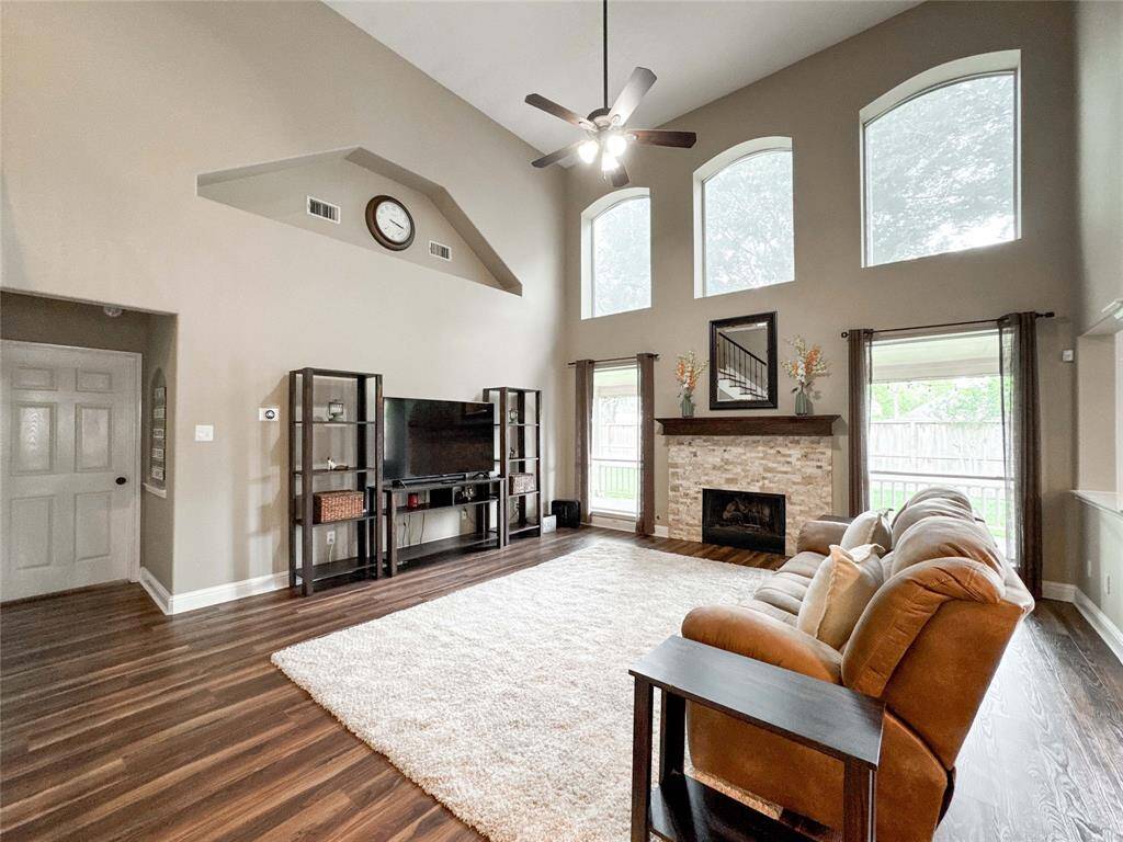 Spacious living room with high ceilings and large windows offering plenty of natural light. Features include a stone fireplace, ceiling fan, and stylish wood flooring. Ideal for comfortable family living and entertaining.