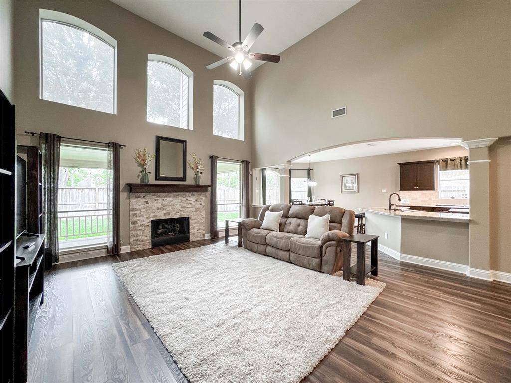 This spacious living area features high ceilings and large windows that provide abundant natural light. It has a cozy fireplace, an open floor plan connecting to the kitchen, and elegant wood flooring, creating an inviting atmosphere.