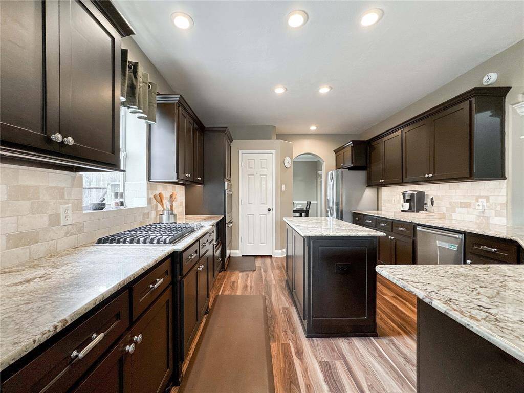 This kitchen features elegant dark wood cabinetry, granite countertops, and a central island. It boasts stainless steel appliances, a gas cooktop, and recessed lighting, all set against a backdrop of warm-toned tile backsplash and wood flooring.