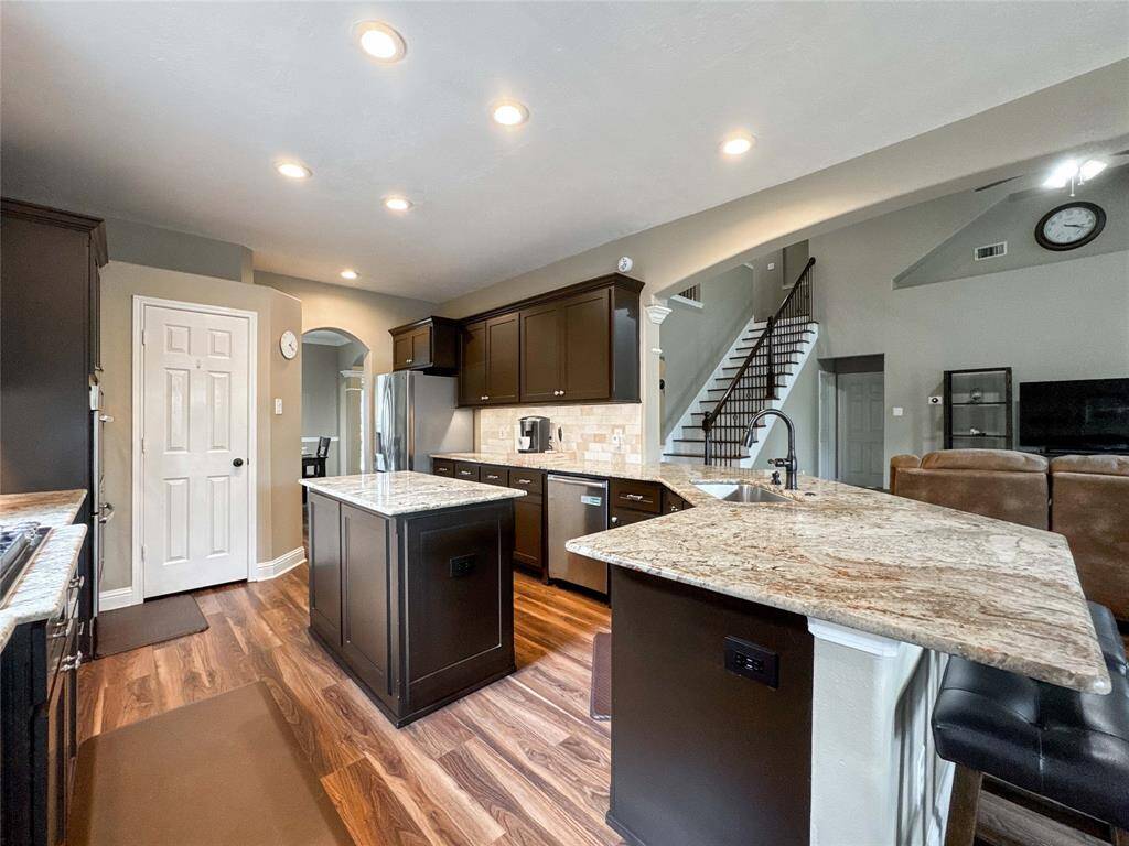 This spacious kitchen features elegant dark cabinetry, granite countertops, and modern stainless steel appliances. The open layout flows seamlessly into the living area, enhanced by warm wood flooring and ample recessed lighting. A stairway leads to the upper level, adding an inviting touch to the space.