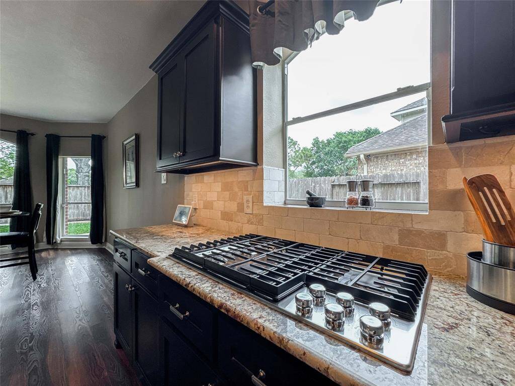 This kitchen features dark cabinetry, a gas cooktop with a sleek granite countertop, and a large window providing ample natural light. The open layout connects to a dining area with hardwood floors and access to the backyard.
