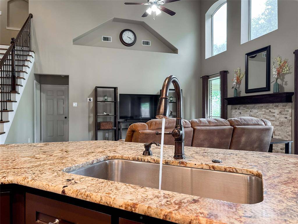 This photo showcases a spacious living area with high ceilings and large windows that provide plenty of natural light. The open layout includes a kitchen island with a granite countertop and modern sink fixture, leading into a cozy seating area with a fireplace and entertainment setup. A staircase adds architectural interest, enhancing the home's inviting atmosphere.