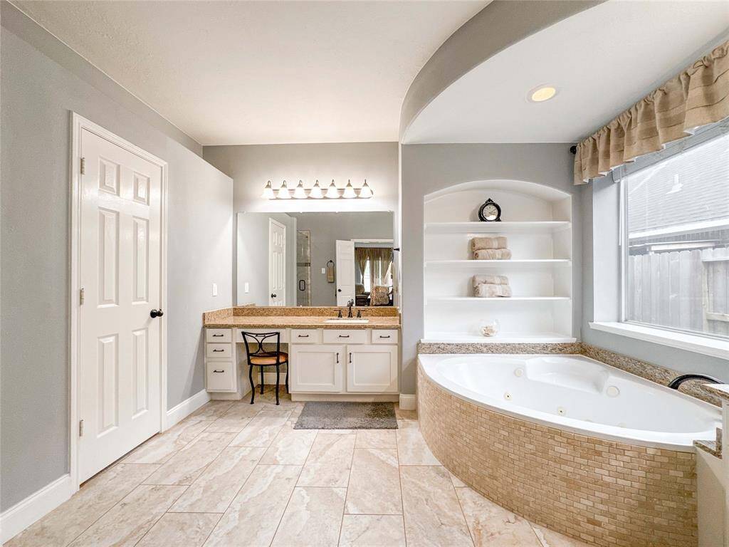 This bathroom features a spacious layout with a large soaking tub, elegant tile flooring, a double vanity with ample storage, and a well-lit mirror. Natural light streams in through a sizable window, enhancing the room's inviting atmosphere.