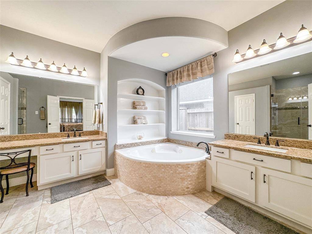 This spacious bathroom features a luxurious corner tub with built-in shelving, dual vanities with granite countertops, and ample lighting. The neutral tile flooring and large window provide a bright and inviting atmosphere.