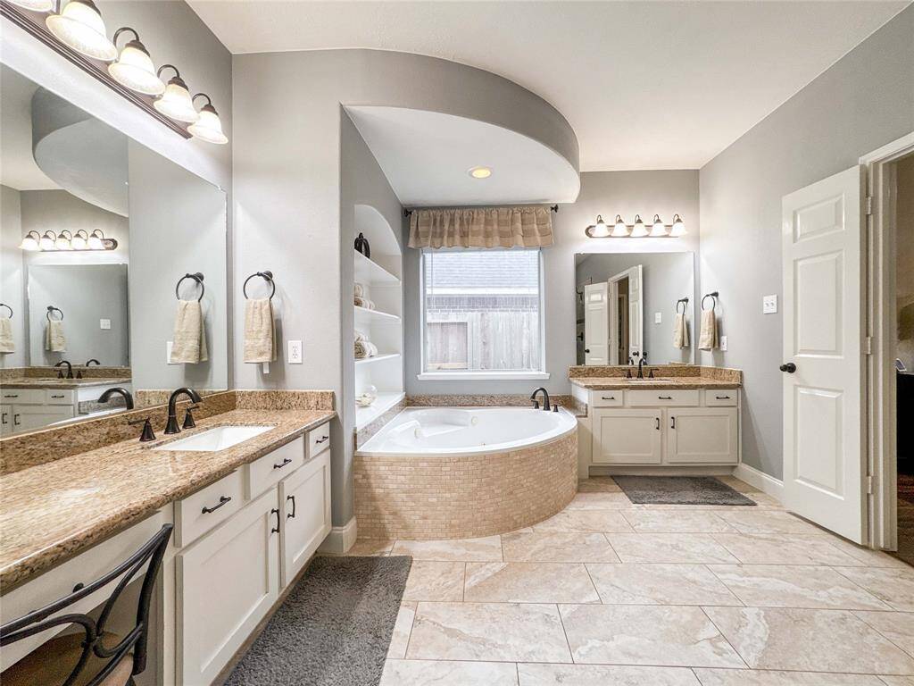 This spacious bathroom features a luxurious corner bathtub, dual sinks with granite countertops, elegant lighting fixtures, and ample storage with built-in shelves and cabinets. The neutral tile flooring and large window provide a bright and inviting atmosphere.
