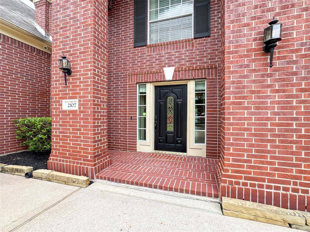 This photo shows a welcoming entrance of a home featuring a sturdy black door with decorative glass panels, surrounded by red brickwork. There are outdoor wall lights on either side, and a small landscaped area with shrubs.