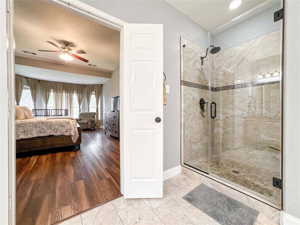 This view showcases a bright primary bedroom with luxury vinyl flooring, a ceiling fan, and large windows with curtains. It connects to an elegant ensuite bathroom featuring a seamless glass-enclosed shower with modern fixtures and tiled walls.