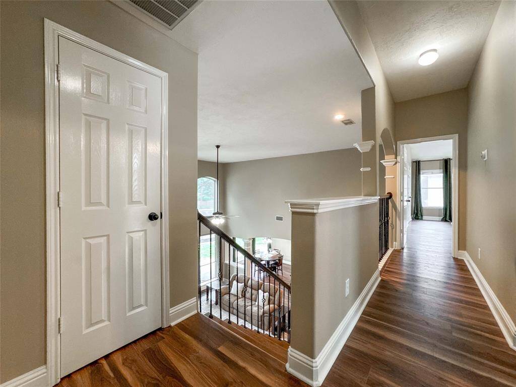 Bright and spacious hallway with luxury vinyl flooring, leading to the game room with large windows. The open staircase with iron railings overlooks a cozy living area below, enhancing the home's open feel.