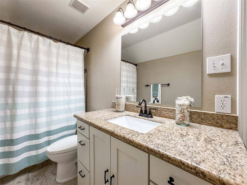 This bathroom features a granite countertop with a single sink, modern fixtures, and ample cabinet space. It has a neutral color palette, a large mirror, and a shower with a striped curtain.