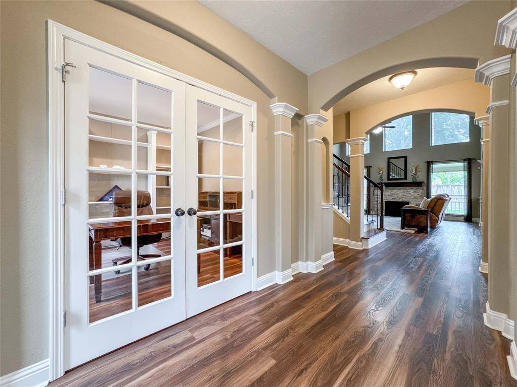 This inviting foyer entry features elegant French doors leading to a cozy office space, arched doorways, and warm wood flooring extending into a spacious living area with a view of large windows and a fireplace.