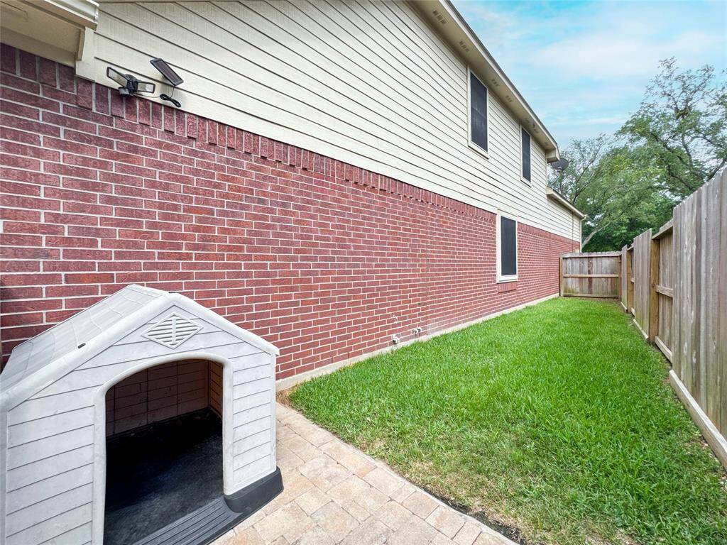 This photo shows a side yard with a brick exterior, a small grassy area, and a paved section with a doghouse. It's bordered by a wooden fence, offering a private and functional outdoor space.
