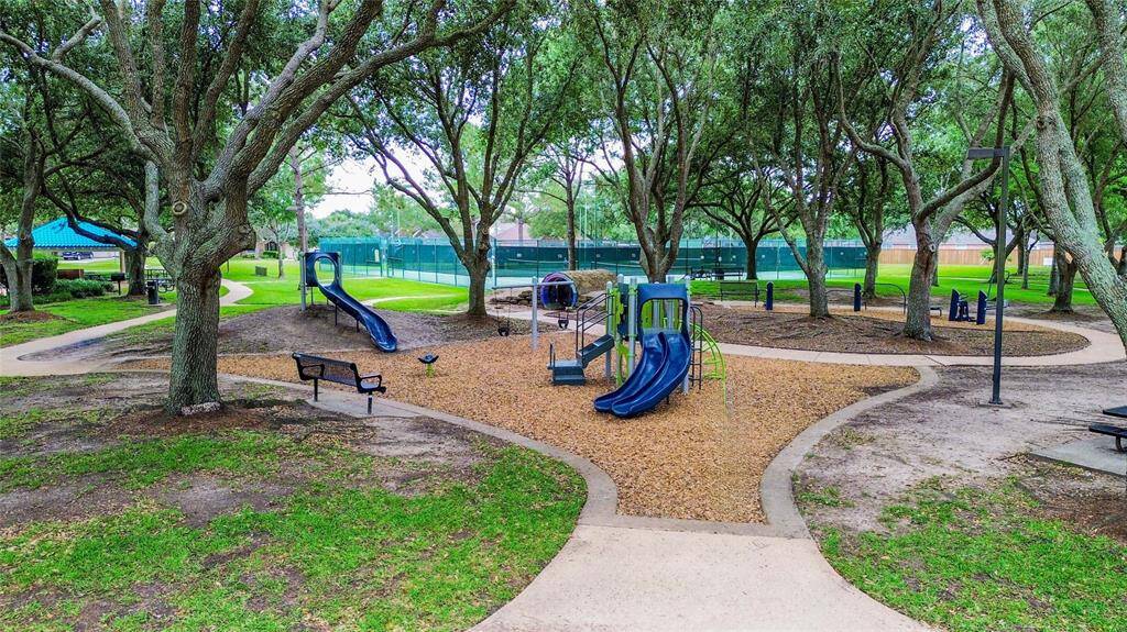 This photo showcases the charming Brittany Lakes Park & Pool area with a playground featuring slides and climbing structures. Surrounded by lush trees, the area offers shaded pathways and benches, making it an inviting space for relaxation.