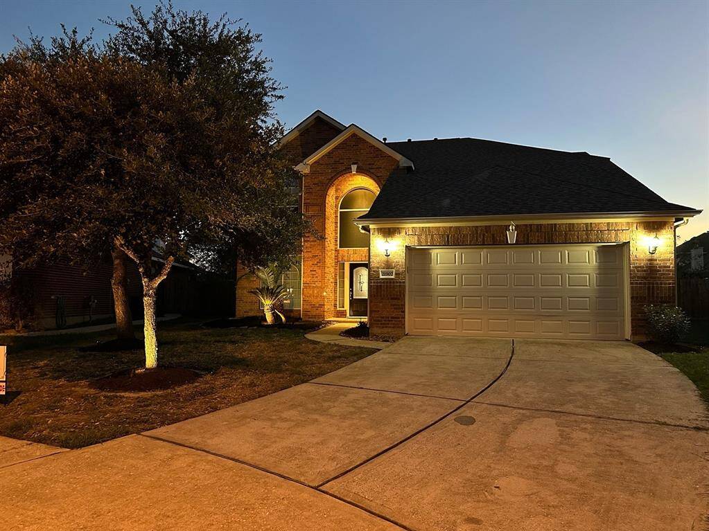 This inviting home features a well-lit brick exterior, a two-car garage, and a neatly landscaped front yard with mature trees, creating a welcoming atmosphere.