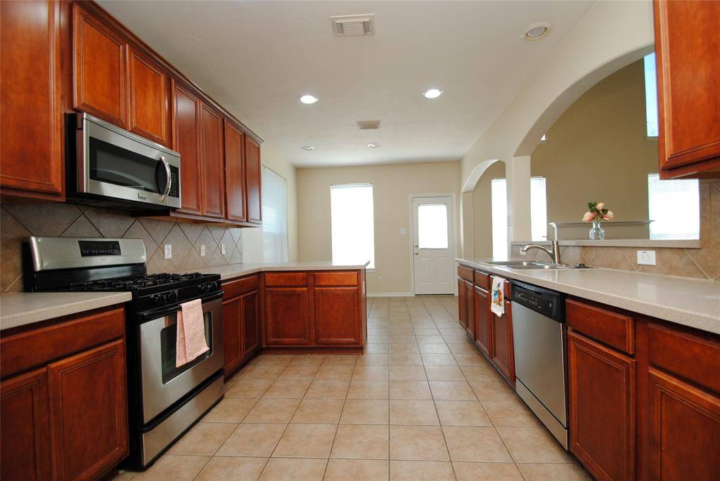 Spacious kitchen with warm wood cabinetry, stainless steel appliances, and ample counter space. Features tiled floors, a dual sink, and a bright, open layout with a pass-through window. Ideal for cooking and entertaining.
