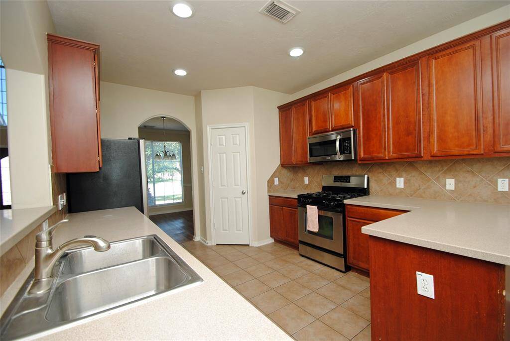 This kitchen features stainless steel appliances, ample wooden cabinetry, and a spacious counter. It has a tiled floor and an archway leading to a dining area with large windows for natural light.