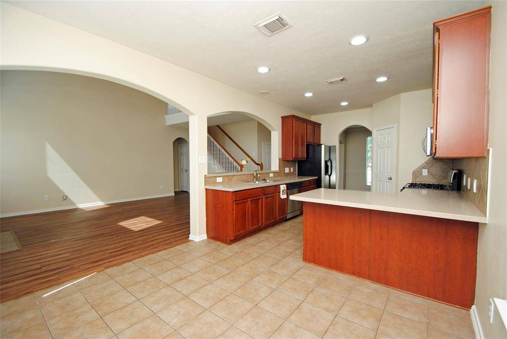 This photo showcases a spacious open-plan kitchen and living area. The kitchen features wooden cabinets, a large countertop, stainless steel appliances, and tile flooring. The living area has wood-look flooring and is well-lit with natural light, creating an inviting space for gatherings.