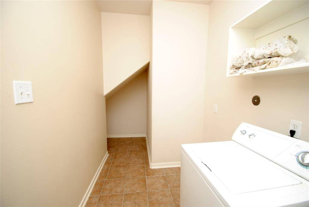 This photo shows a compact laundry room with tiled flooring, featuring a washing machine and an upper shelf for storage. The room has neutral-colored walls and a small alcove space for additional storage.