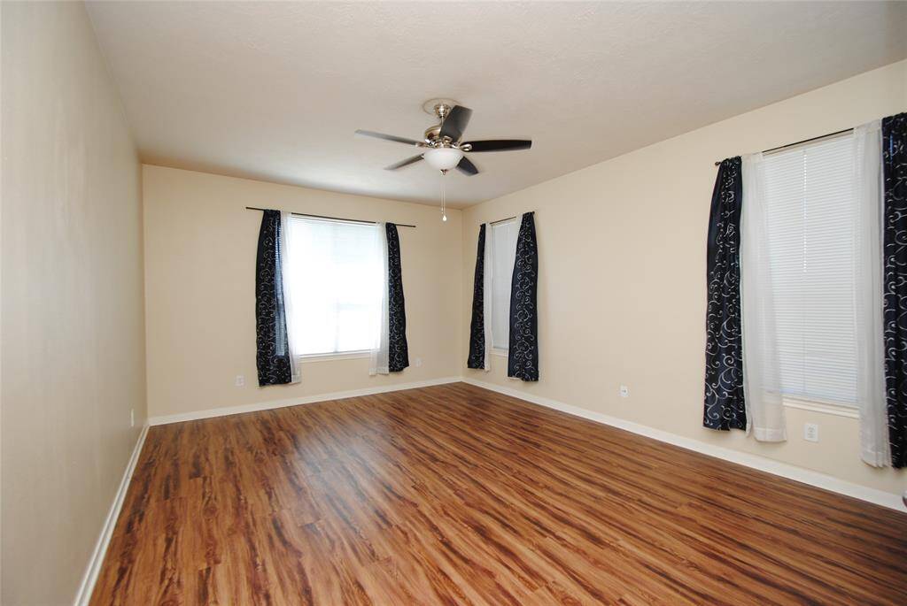 Bright, spacious room with wood laminate flooring, neutral walls, and large windows adorned with elegant dark curtains. A ceiling fan provides comfort and style.