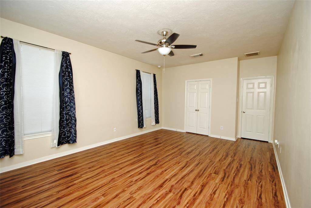 This room features laminate wood flooring, neutral walls, and a ceiling fan. It has two doors and two windows with dark, patterned curtains, offering a spacious and versatile area.