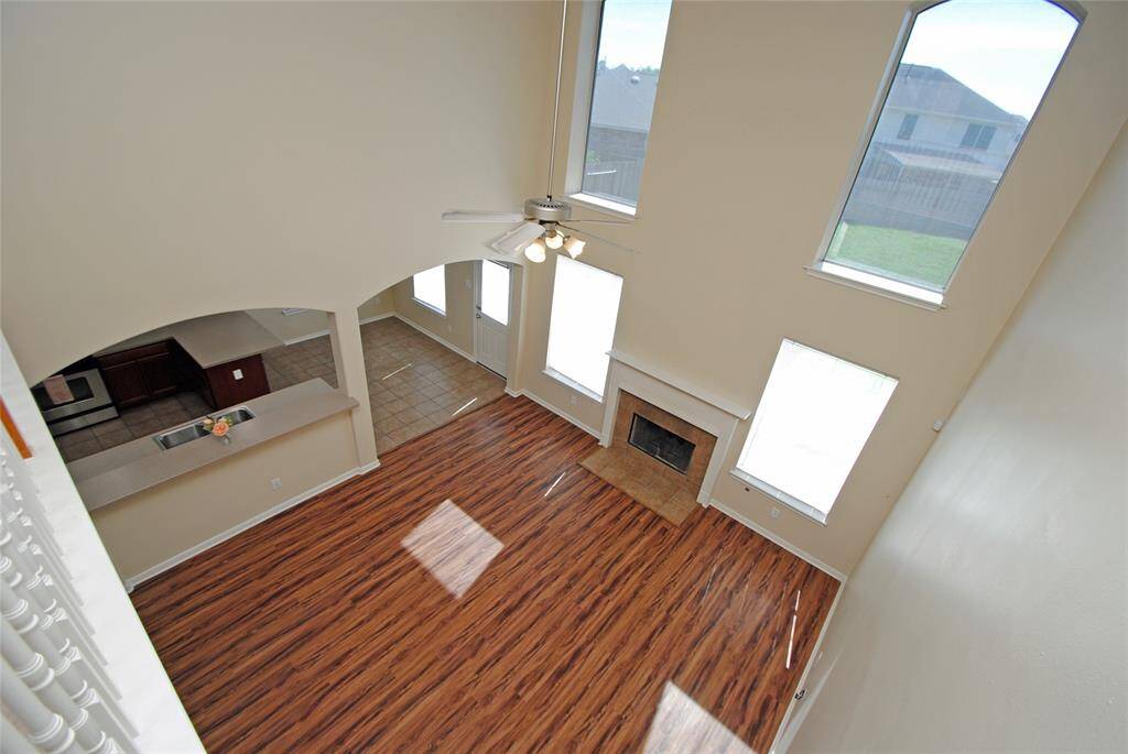 This spacious living room features high ceilings, large windows for natural light, a cozy fireplace, and wood-style flooring, with an open view of the adjacent kitchen area.