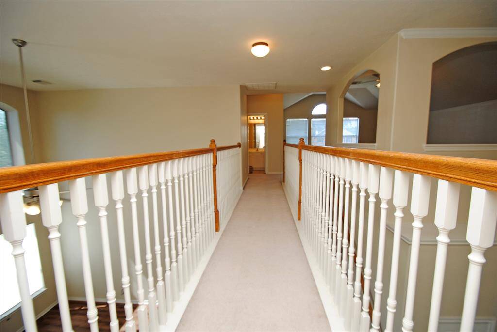 This photo showcases an upstairs hallway with a carpeted floor and wooden railing. It leads to a well-lit space with large windows, creating an open and airy feel. Arched doorways add an elegant touch to the design.