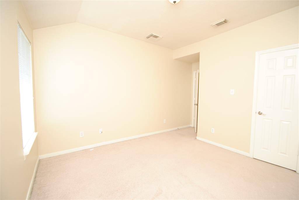 This is a bright, empty room with neutral beige walls and carpet. It features a window for natural light, a standard ceiling light, and a white door. Ideal for a bedroom or office space.