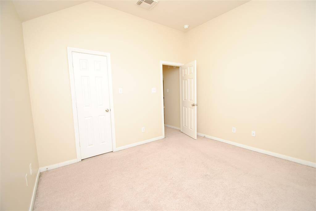 This is a small, empty room with beige walls and carpet. It features a closed closet door and an open interior door, with a high ceiling and soft lighting.