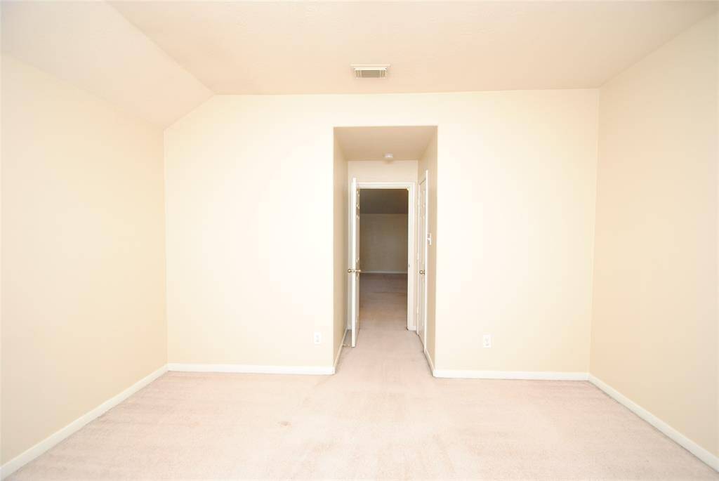 This photo shows a neutral-toned room with a vaulted ceiling and carpet flooring, leading to a hallway through a door, providing a spacious and versatile living area.