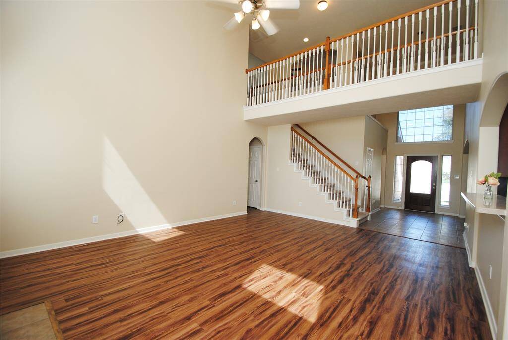 This spacious living area features high ceilings, a wooden floor, and an elegant staircase leading to an upper level. Natural light fills the room through large windows, and the entrance showcases a grand doorway with tiled flooring. Perfect for open-concept living.