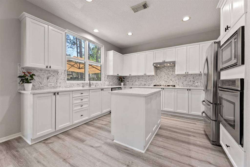 This kitchen features modern white cabinetry, stainless steel appliances, and a central island. It has a sleek marble-patterned backsplash and ample natural light from large windows, complemented by light wood flooring.