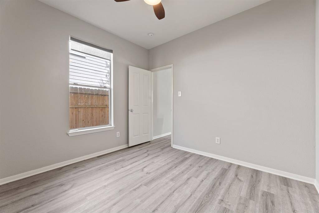 Study features light gray walls, wood-look flooring, and a ceiling fan. It has a window with natural light and a view of a wooden fence, creating a cozy, neutral space.