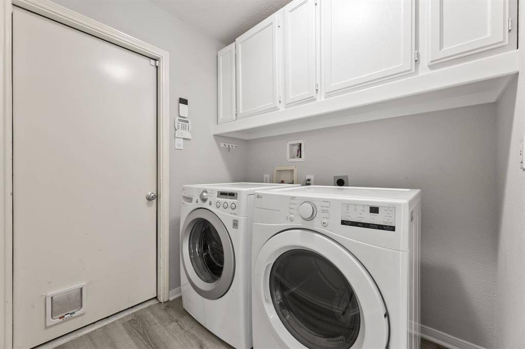 This photo shows a modern laundry room with a front-loading washer and dryer. It features white cabinetry above for storage, a light color scheme, and a door with a pet door installed. The space is compact and functional.