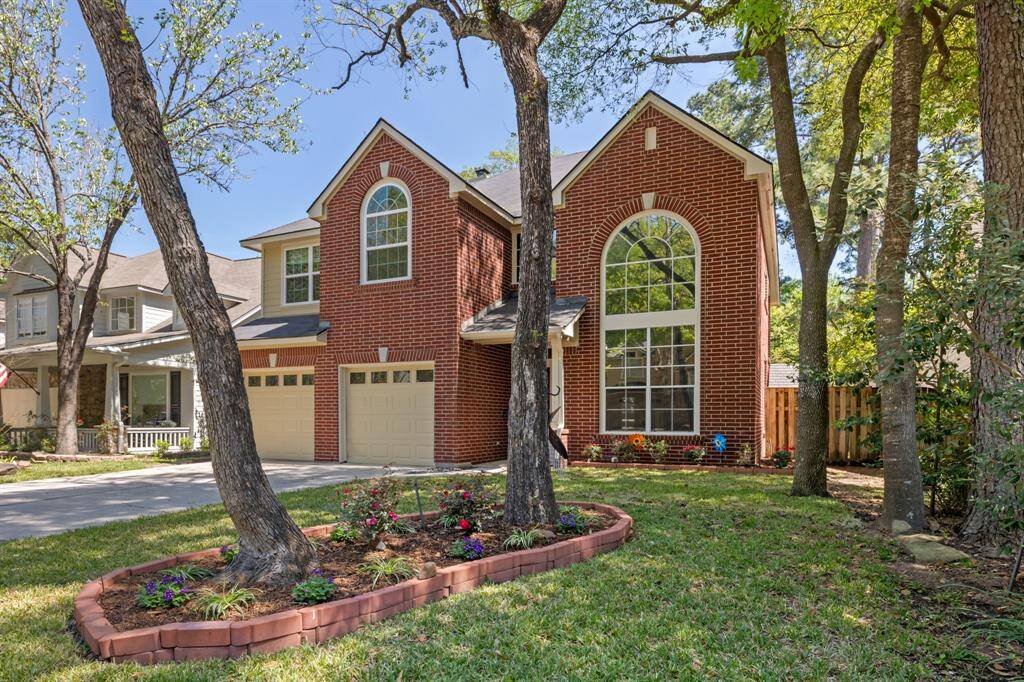 Charming two-story brick home featuring a large arched window, two-car garage, and a well-maintained front yard with mature trees and landscaped garden beds.