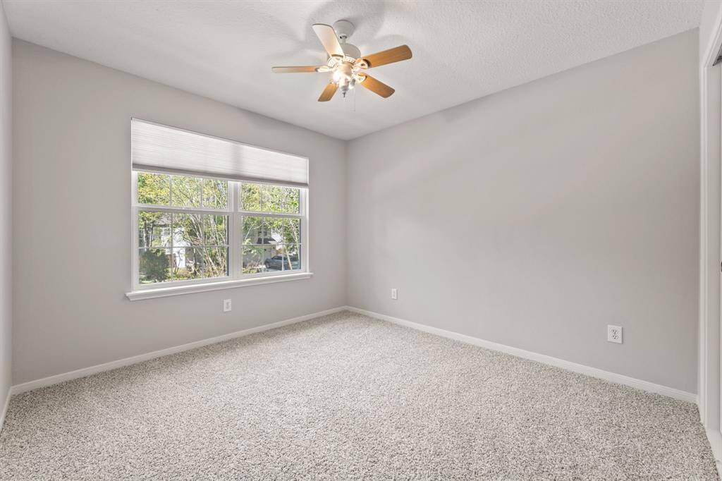 This is a bright, carpeted bedroom with neutral walls, a large window offering natural light, and a ceiling fan.