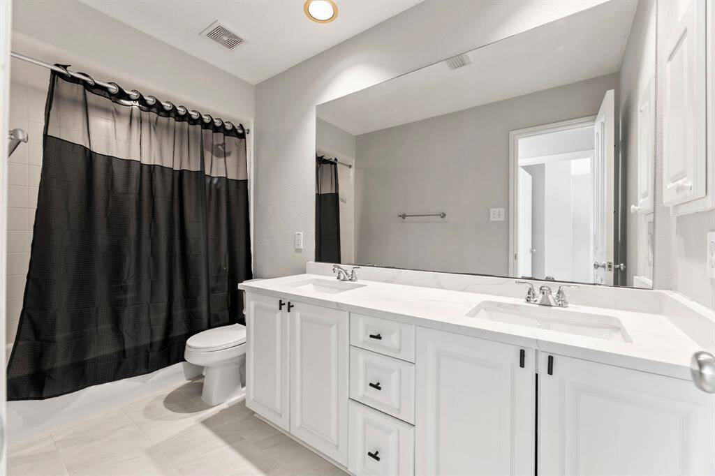 This bathroom features a clean, modern design with a dual-sink vanity, ample storage, and a large mirror. The space is bright with neutral tones, complemented by a shower with a dark curtain.