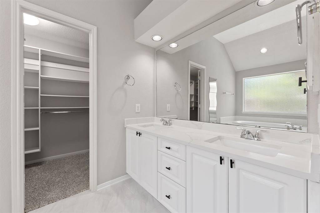 This spacious Primary bathroom features a dual-sink vanity with ample storage and modern fixtures. It includes a large mirror, recessed lighting, and an adjoining walk-in closet with built-in shelving. A frosted window allows natural light while maintaining privacy.
