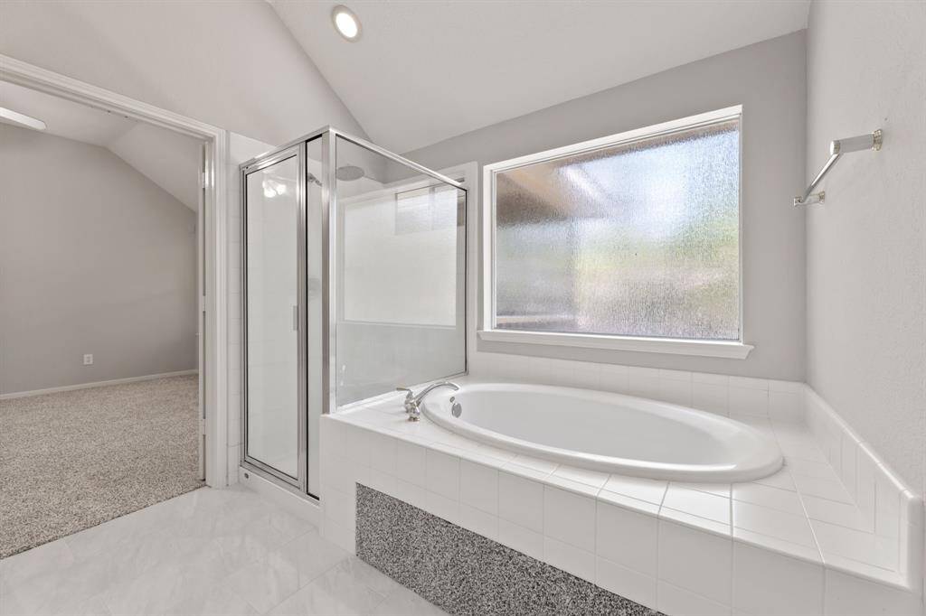 This Primary bathroom features a spacious bathtub with white tile surround, a separate glass-enclosed shower, and a large frosted window for natural light and privacy. The neutral color palette creates a clean, modern look.