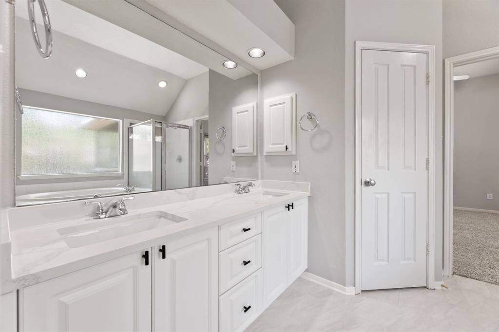 This bright Primary bathroom features dual sinks with modern fixtures, a spacious countertop, and ample storage with white cabinetry. It includes a walk-in shower and a large frosted window for natural light.