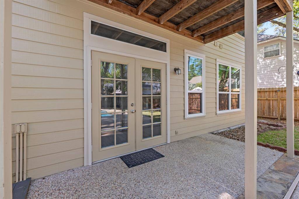 This photo showcases a cozy backyard patio with French doors, a covered area with wood paneling, and adjacent windows allowing natural light. The space is bordered by a wooden fence, providing privacy and a view of a lush garden area.