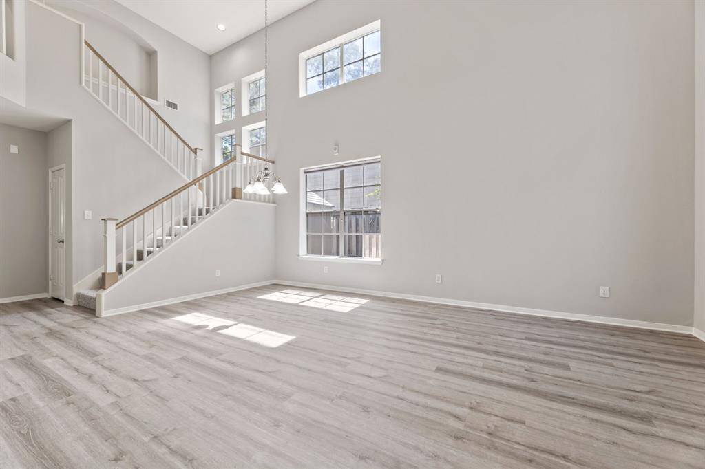 This spacious living area features high ceilings, abundant natural light from tall windows, and modern light wood flooring. A stylish staircase adds architectural interest, leading to an upper level.
