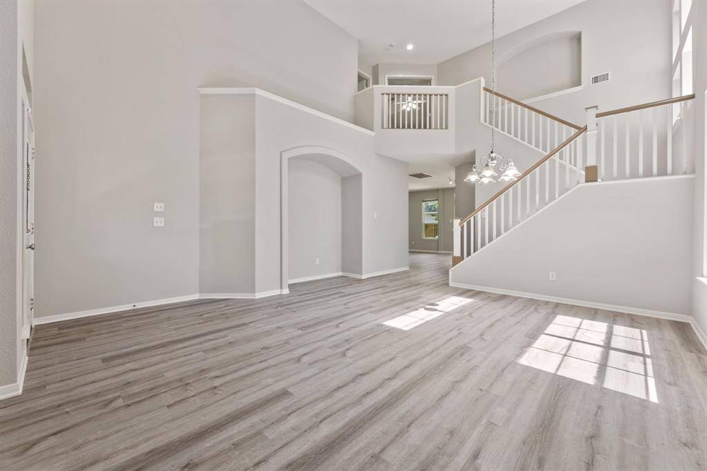 This spacious, light-filled living area features high ceilings, modern gray laminate flooring, and a stylish staircase leading to an upper level. The open layout is complemented by large windows and a contemporary chandelier, creating an inviting atmosphere perfect for any homebuyer.