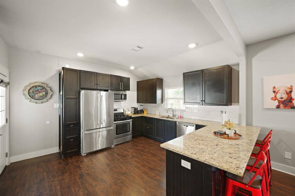 This cozy kitchen features modern gray cabinetry, stainless steel appliances, and a compact dining area with a wooden table and bar stools. The space is well-lit with recessed lighting, and a large wall clock adds a stylish touch.