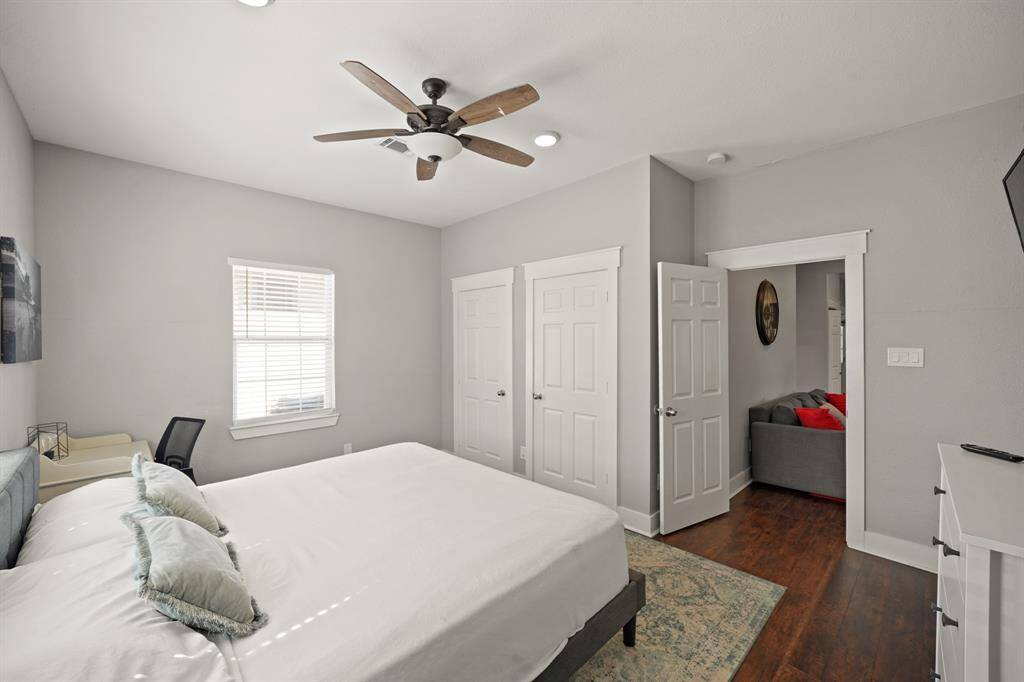 This bedroom features a neutral color palette with soft gray walls and a cozy bed. It includes a wall-mounted TV, a small dresser with decorative items, and a closet with white double doors. A colorful artwork adds a touch of vibrancy. The room is well-lit and inviting.