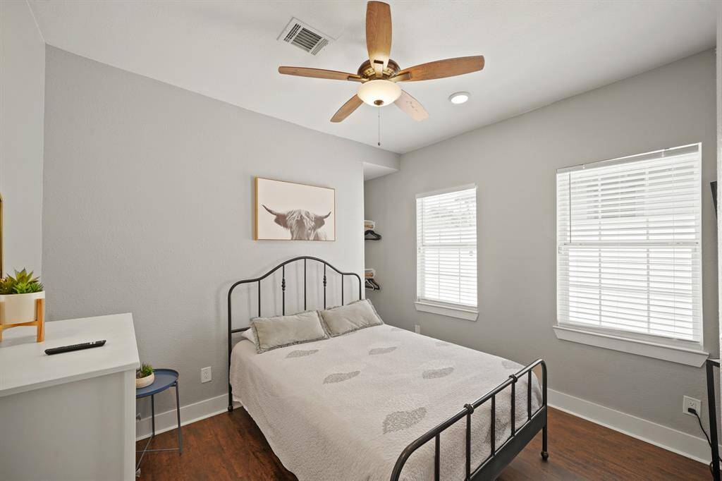 This bedroom features a cozy atmosphere with a metal-framed bed, hardwood floors, and neutral walls. It includes a wall-mounted TV, a white dresser, and two windows for natural light.