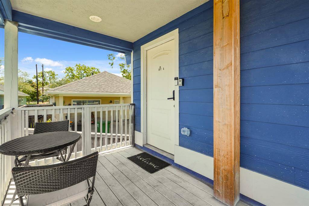 charming porch area with blue siding, featuring a welcoming entrance door. The space includes a small outdoor seating area with a round table and chairs, ideal for relaxation. The porch is covered, offering shade, and overlooks a neighboring house with a well-maintained roof, surrounded by greenery.