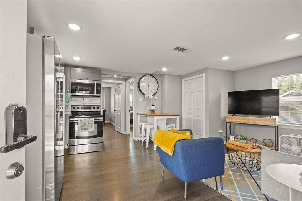 inviting living area features modern decor with a cozy seating arrangement, a wall-mounted TV, and a small dining nook. The open layout connects to the kitchen, and natural light fills the space through a window, highlighting the stylish hardwood like floors.