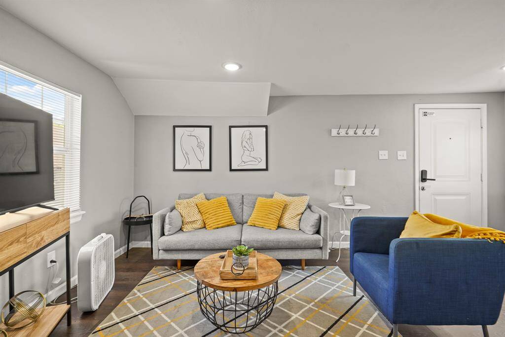 This cozy interior features a compact kitchen area with modern appliances, a wood-topped island with seating, and a stylish wall clock. The space is accented by hardwood floors and neutral walls, leading to multiple rooms and a small TV stand.